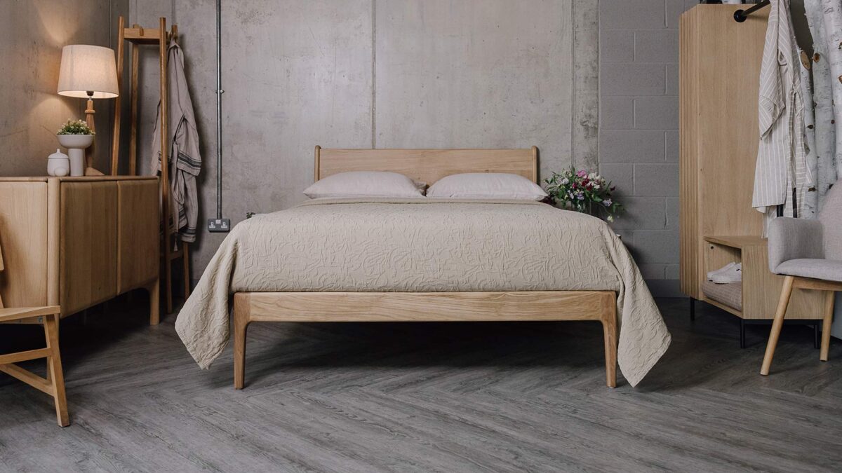 Zoomed-out shot of the English oak Camden, showing its headboard and foot-rail. It is dressed with the floral textured bedspreads in the ecru colour-way. On one side of the bed sits the Ethnicraft oak Pi sideboard and the Tower oak coat-stand, and on the other side of the bed sits the Modern Oak Open Wardrobe.