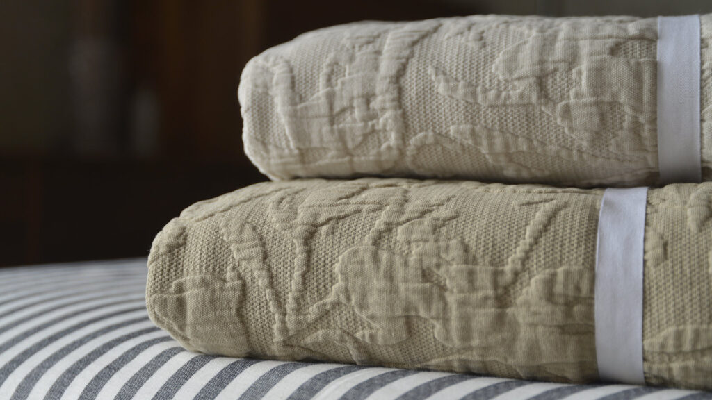 Folded stack of floral textured bedspreads, showing the light ecru colour-way on the camel colour-way, sat atop black and white striped bed linen.