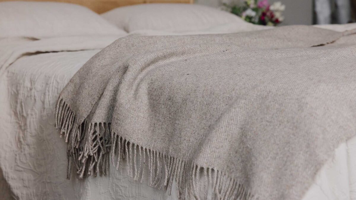 Greige lambswool merino throw with tassels draped across a bed.