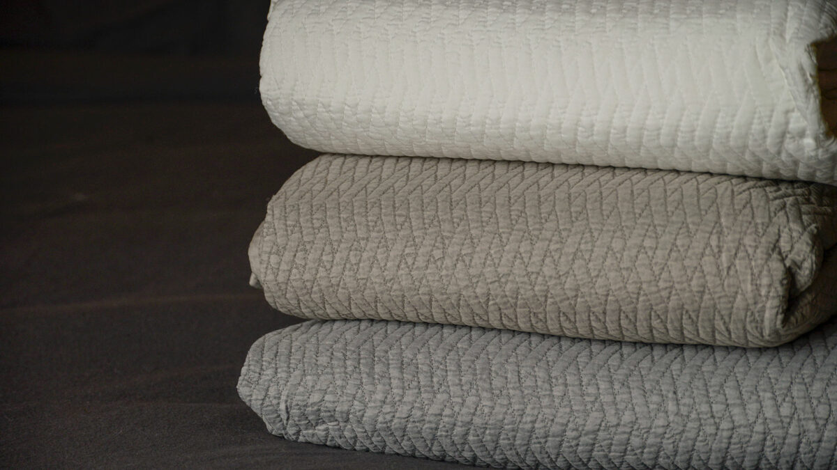 stitched design quilted bedspread in white, taupe, or grey shown folded in a stack