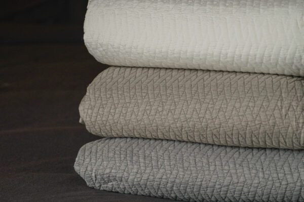 stitched design quilted bedspread in white, taupe, or grey shown folded in a stack