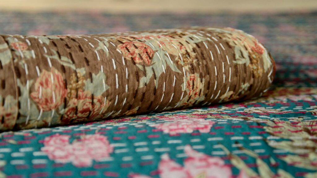 folded brown kantha throw with floral pattern and brown and white stitching sitting on the teal version of the same throw.