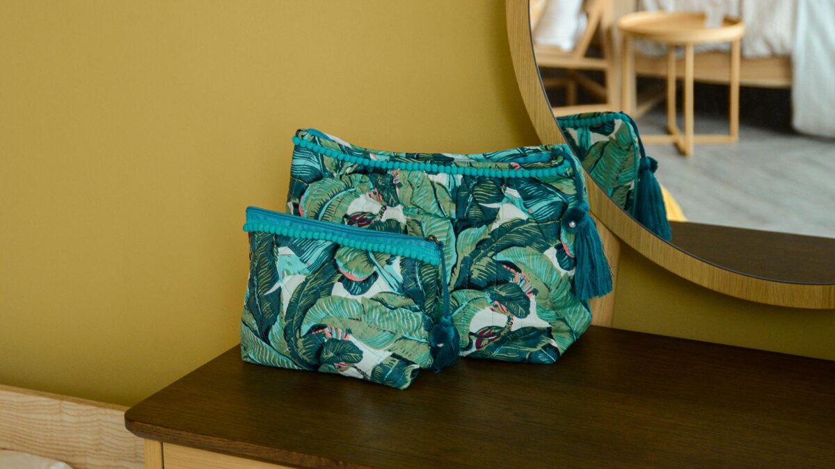 Two blue and green tropical print toiletry bag with large tassels, one small, one large, sat on dark table top with mirror in the background.