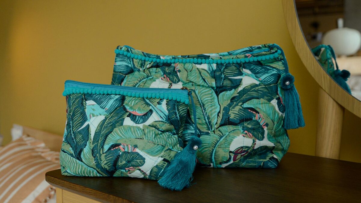 Two blue and green tropical print toiletry bag with large tassels, one small, one large, sat on dark table top with mirror in the background.