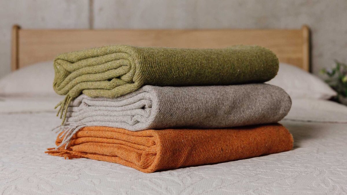 Stack of three lambs wool throws on a bed. Top to bottom - Green, griege, burnt orange.