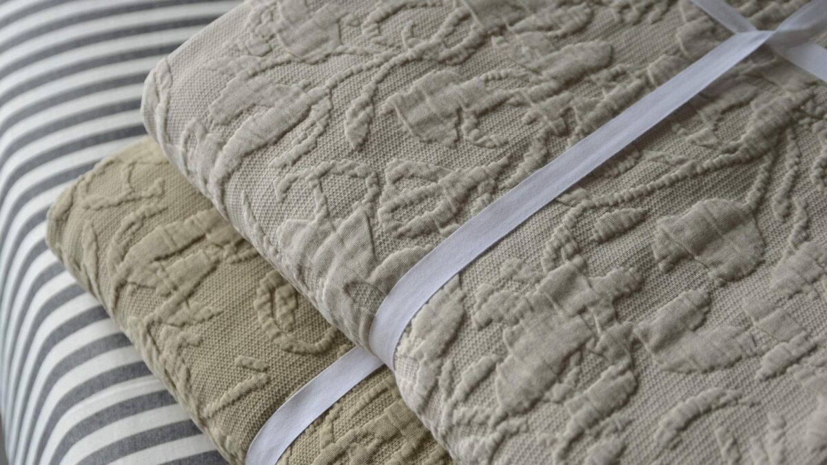 Top view of a stack of folded floral textured bedspreads, showing the light ecru colour-way sat on top of the sand colour-way.