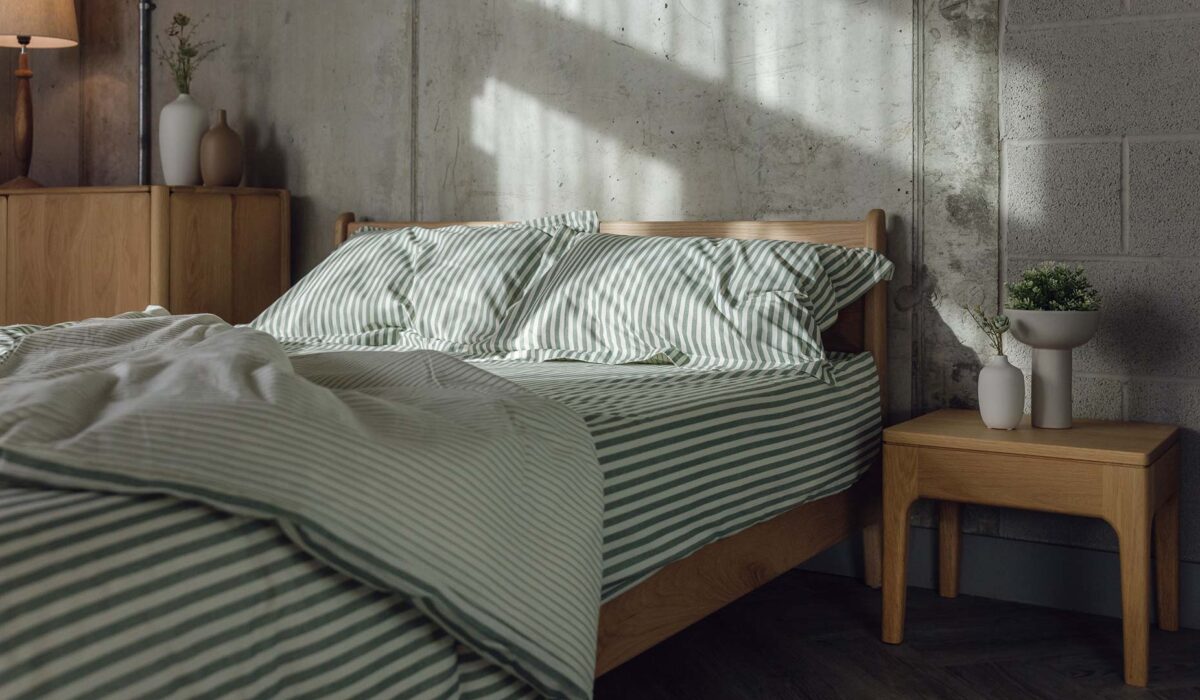 Low oak Pimlico bed base with rumpled green and ivory stripes bedding. Pictured with the original Camden table in oak with vases of greenery.