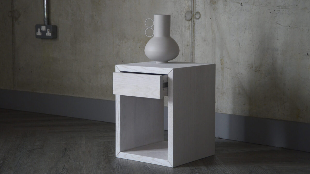 Image showing a small bedside table stained in a pure white finish. A decorative vase sits atop it, and the drawer featured in the table is slightly open.