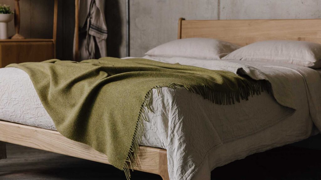 Olive green merino throw with tassels on top of beige linen on an English Oak Camden Bed Frame.