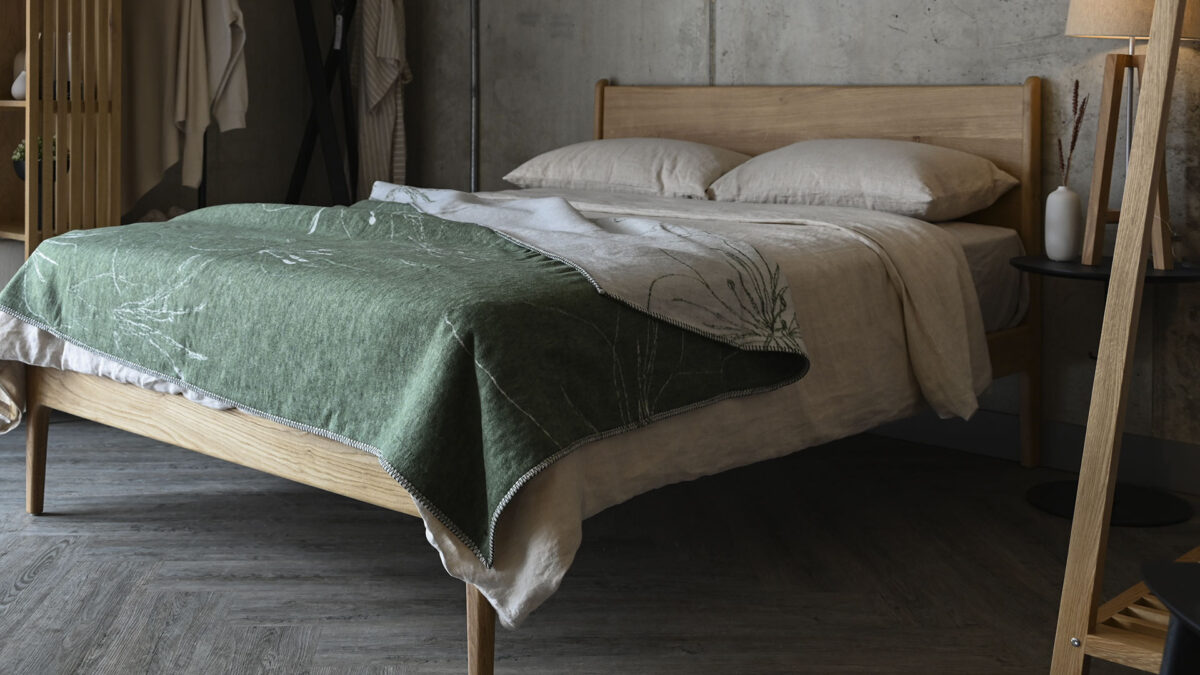 Oak bed with neutral linen and an olive green and ivory floral patterned throw on the top.