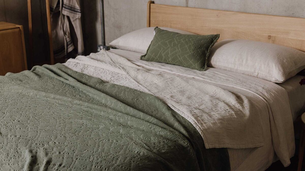 Top down, side view of English Oak bed with beige bedding, topped with an olive green textured pillow and throw.