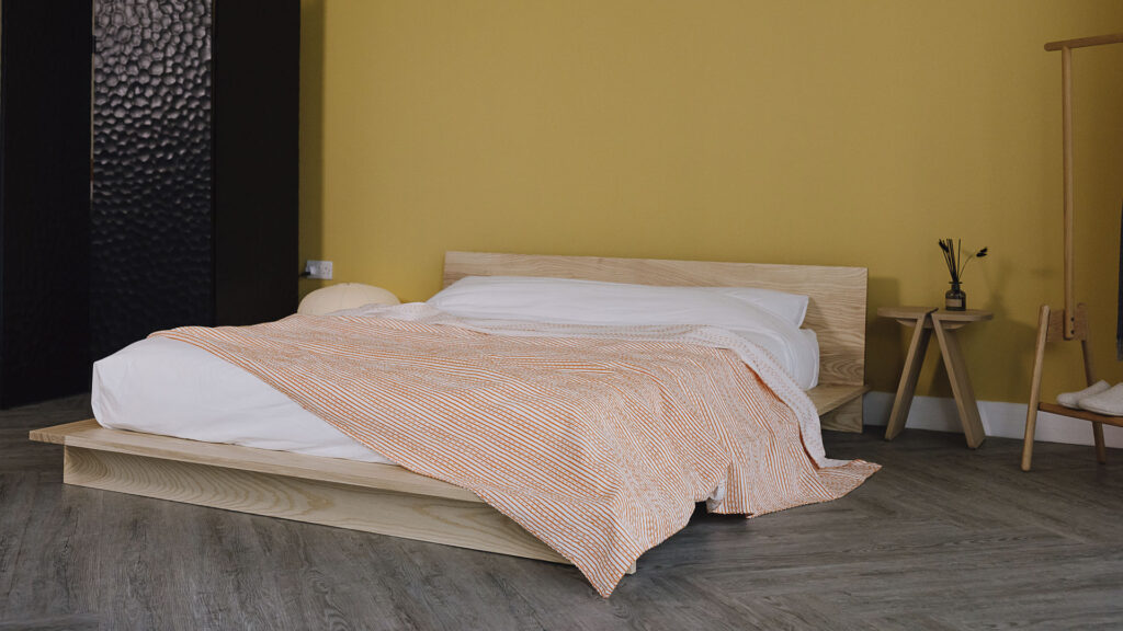 Image of a bedroom scene with a yellow wall and ash bed. Focus of the photo the orange striped bedspread laid over the bed.