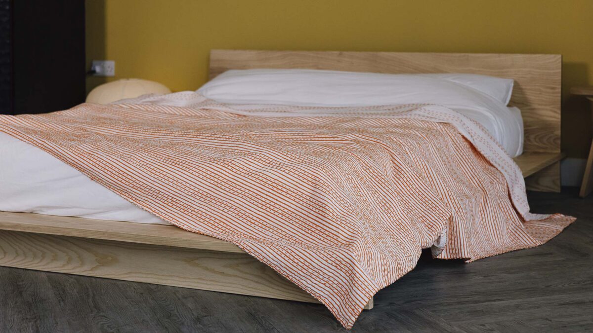 3/4 angle photo. Orange Striped bedspread draped over white bed linen on a ash platform bed frame.