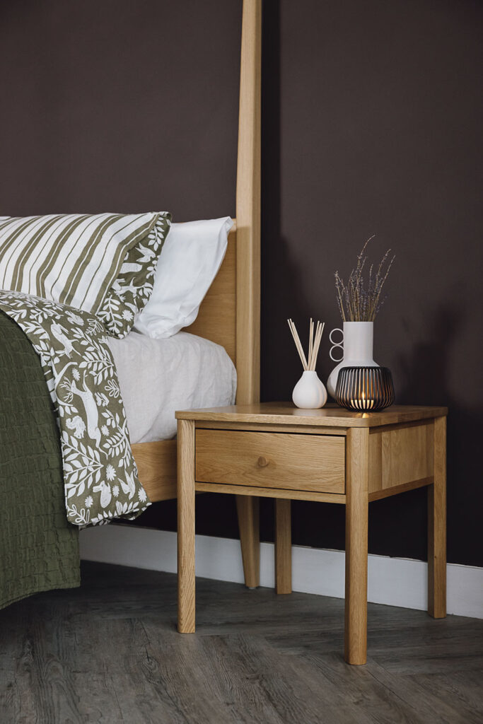 Oak bedside table with vase, diffuser and candle holder next to an oak bed with green patterned linen.