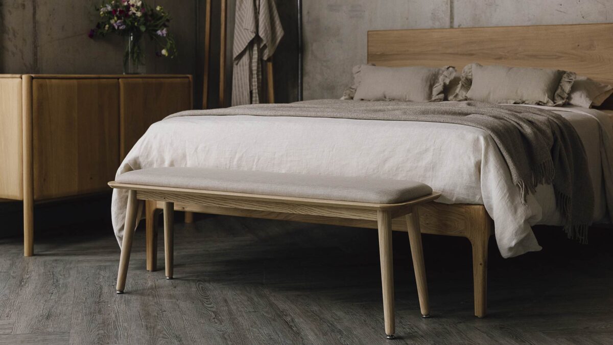 Padded bench at the foot of an oak bed with ecru bedding. Oak sideboard and coat stand in the background.