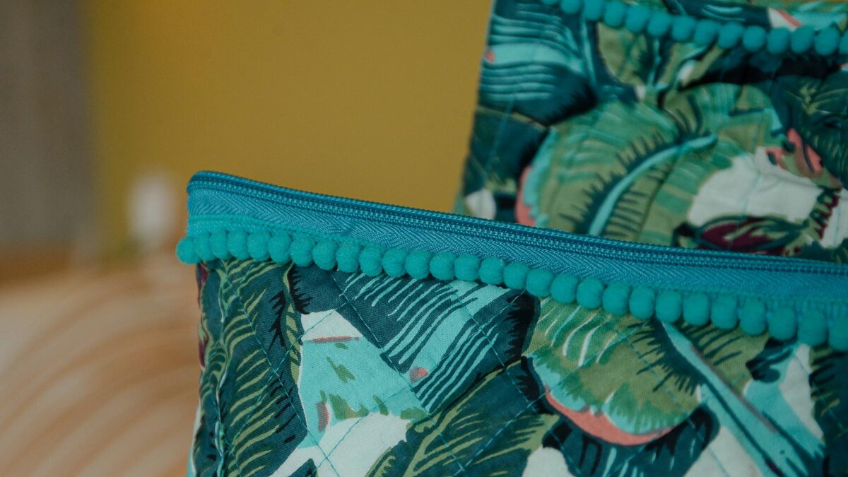 Close up of zip on green and blue tropical print toiletry bags highlighting pom pom detailing.