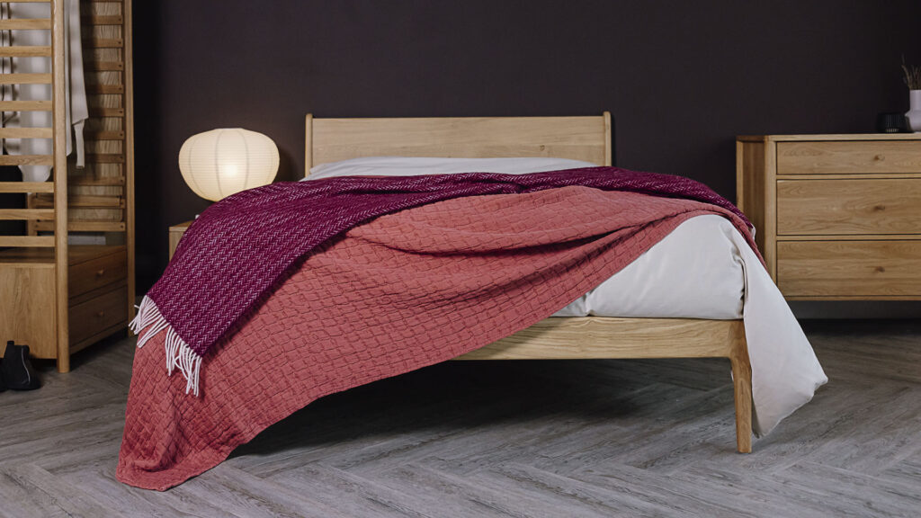 Bed scene with oak furniture. Bed dressed with off neutral linen, topped with a raspberry textured bedspread and a deep burgundy herringbone throw.