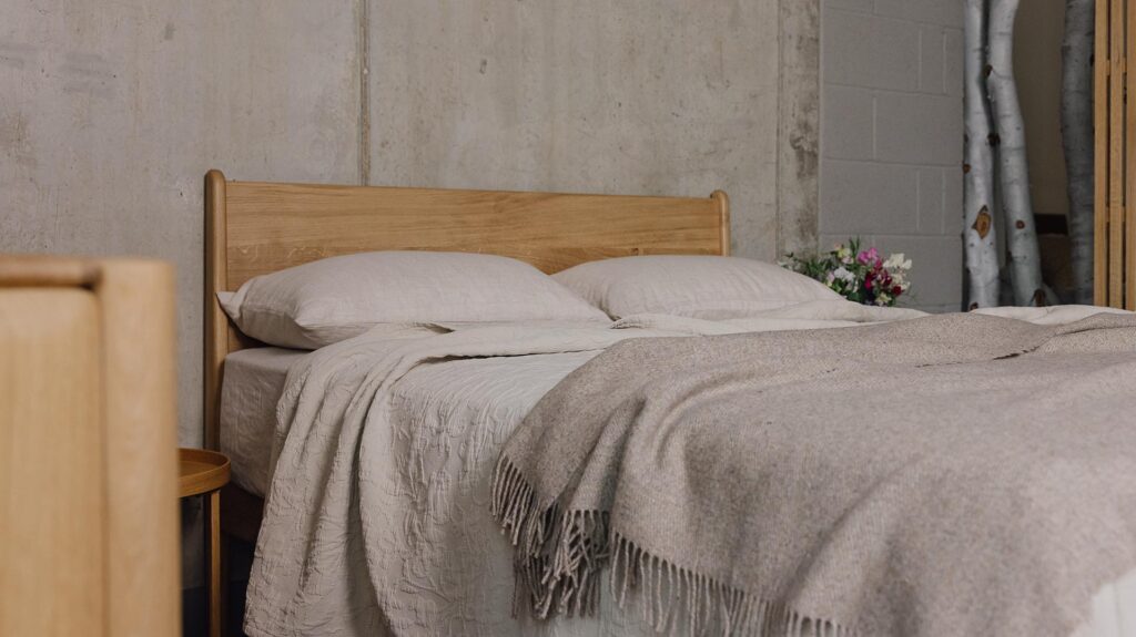 3/4 angle photo of an oak bed with stone linen and a greige lambswool throw.