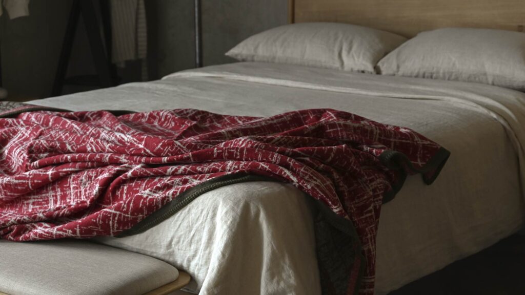 Red throw with white crosshatch pattern and Olive border messily throw over a bed with stone linen.
