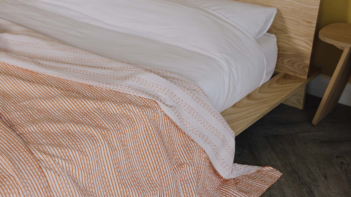 Ash Oregon bed frame with white linen. Orange stripy kantha bedspread on the top, top folded down so that orange stitching detail is visible on the reverse.