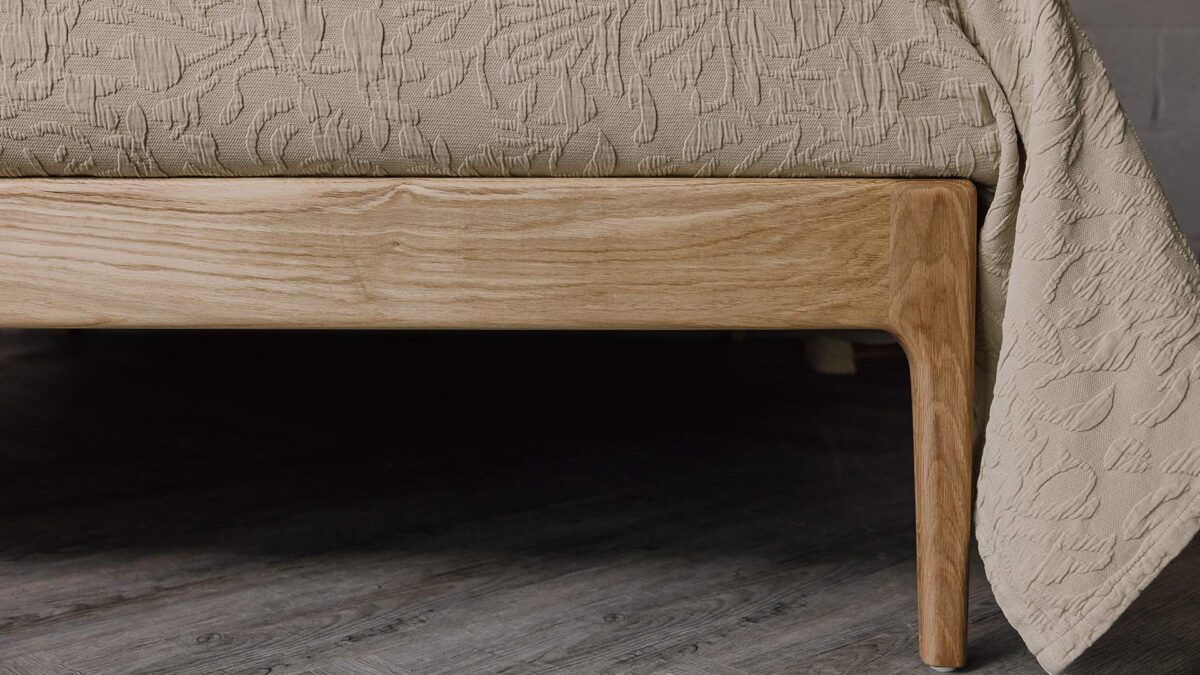 Close up of the English oak Camden's foot-rail. The bed is dressed in the sand-coloured floral textured bedspread.