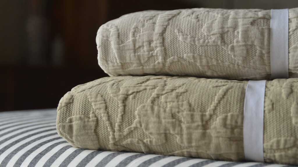 Stack of textured floral cotton bedspreads, in light ecru (on the top) and sand (on the bottom), sat atop black and white striped bed linen.