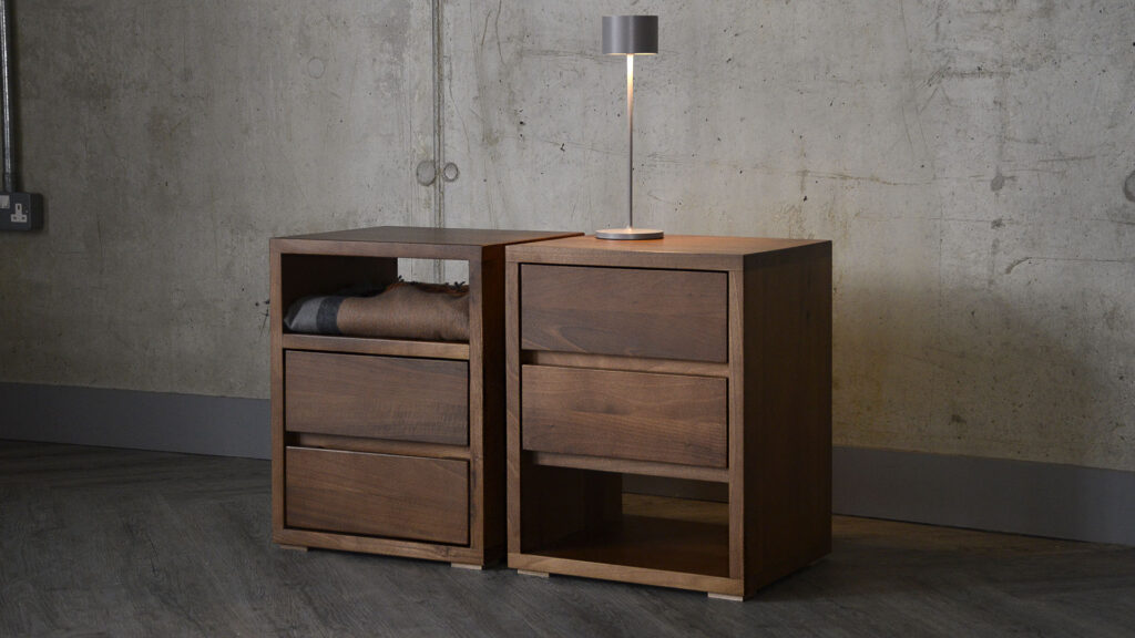 Two Black Lotus Cube bedside tables in walnut sat side by side. Image shows the two options available - either shelf at top or shelf at bottom. The unit to the right has a rechargeable lamp sat atop it, and the unit to the left has a throws folded on the top shelf.