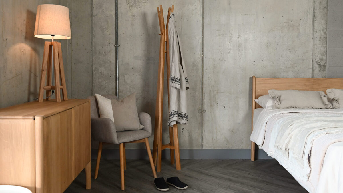 Spire hat and coat stand in modern room setting. The coat stand has a cotton robe hanging from it, and stands next to a grey upholstered chair with a cushion on it, as well as a modern oak sideboard. The image also features the Camden bed base in English Oak to the right hand side of the image.