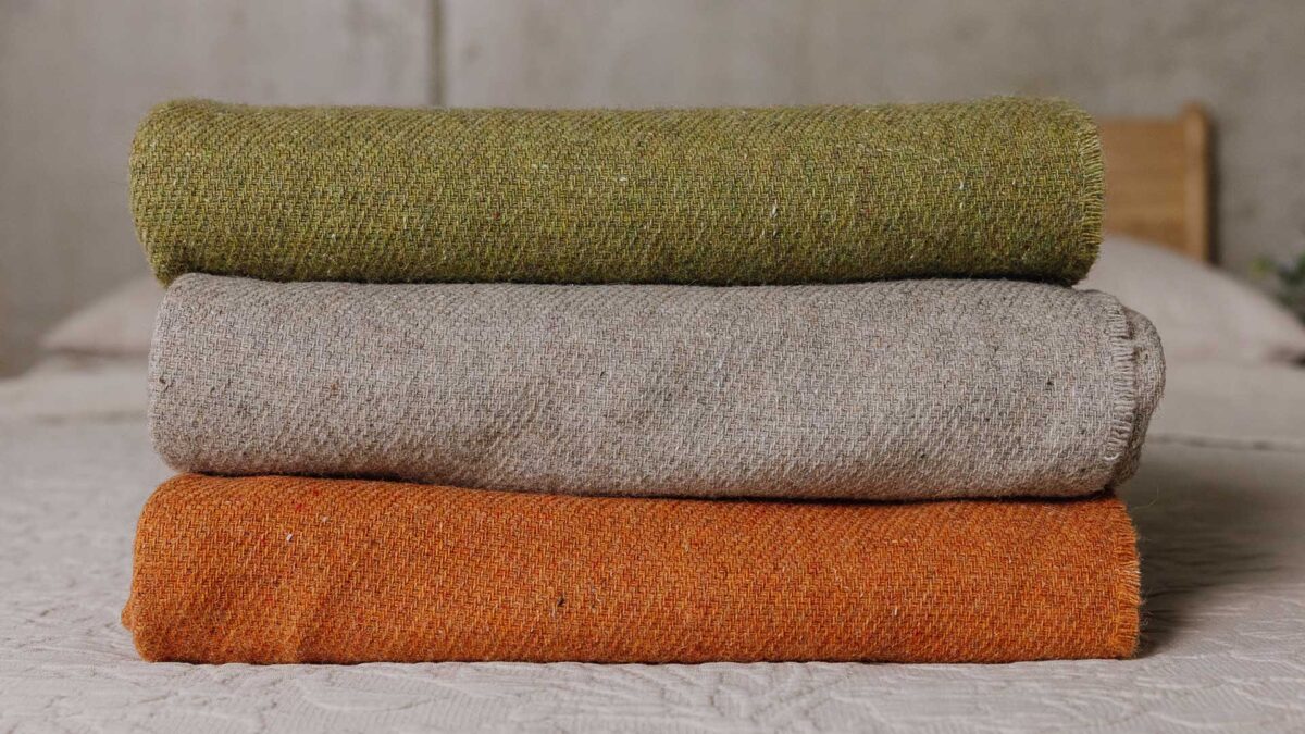 Stack of three lambswool throws on a bed. Top to bottom - Green, griege, burnt orange.
