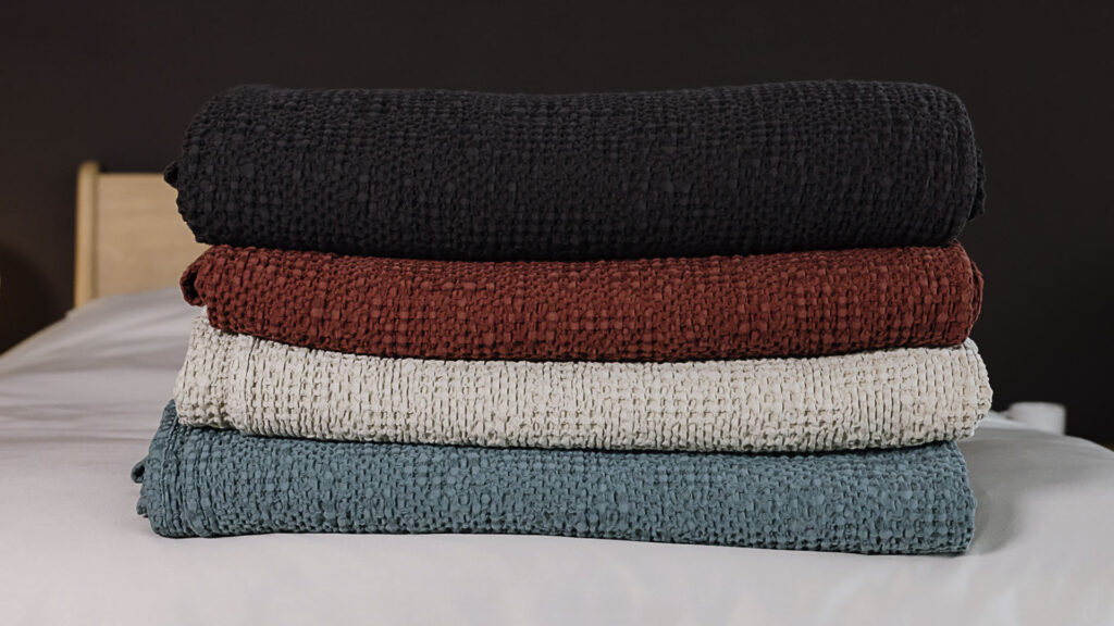 Stack of 4 waffle textured bedspreads neatly folded on a bed. From bottom to top, black, red, ecru and green.