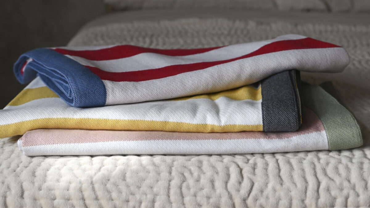 3 striped cotton blankets neatly folded on a bedspread. Bottom to top; Pink and white stripes with a green border, yellow and white stripes with a dark grey border, white and red stripes with mid-blue border.