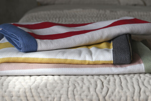 3 striped cotton blankets neatly folded on a bedspread. Bottom to top; Pink and white stripes with a green border, yellow and white stripes with a dark grey border, white and red stripes with mid-blue border.