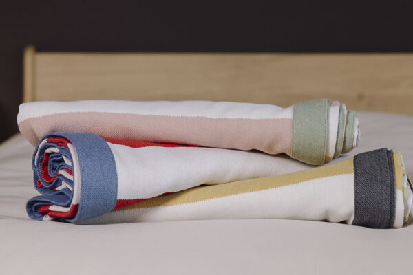 Three striped throws with contrasting border rolled on bed. From bottom to top, yellow and white with grey border, white and red with blue border, pink and white with sage green border.