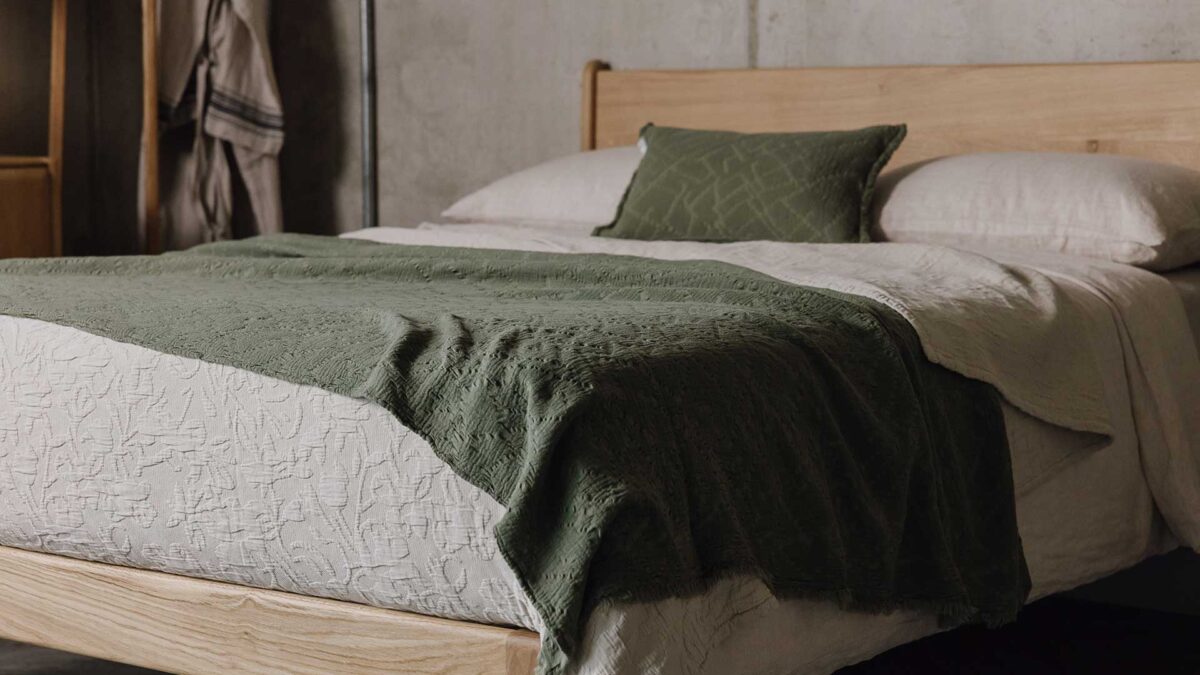 English oak Camden bed frame with beige bedding and an olive green throw and pillow. Side angle, bottom corner of the bed in focus.