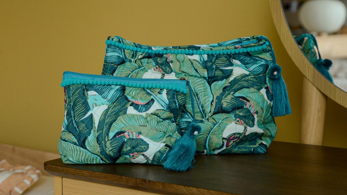 Two blue and green tropical print toiletry bag with large tassels, one small, one large, sat on dark table top with mirror in the background.