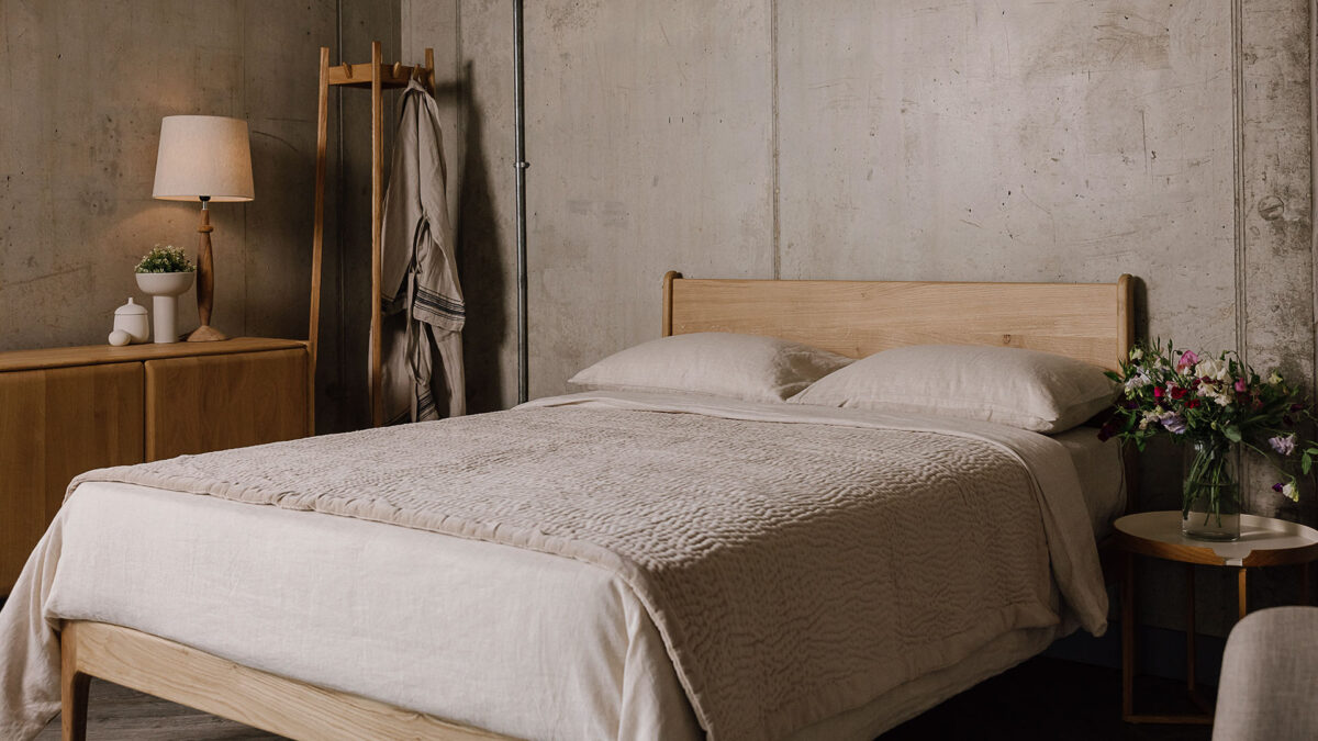 Side on view of the English oak Camden, showing its headboard and part of the foot-rail. The bed is dressed in the organic linen bedding in ecru, and the stitched velvet bedspread in soft beige. There is a round-top bedside table next to it, with clear vase full of colourful flowers on top.