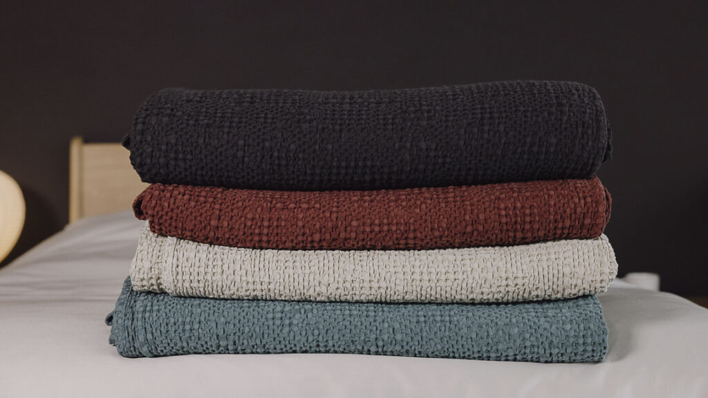 Stack of 4 waffle textured bedspreads neatly folded on a bed. From bottom to top, black, red, ecru and green.