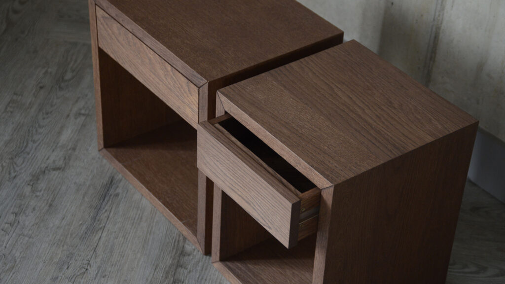 Image shows two bedside tables with a dark brown, walnut-look finish. One of the bedside tables has a square tabletop, whereas the other is wider, with a rectangular tabletop.