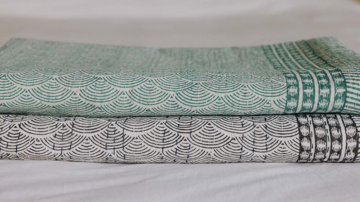 stack of two neatly folded block print bedspreads, top one with a green shell print, bottom grey.