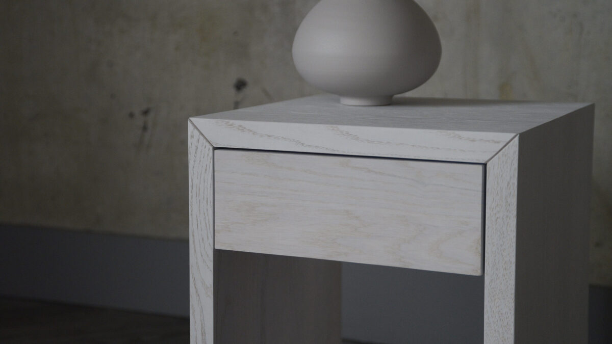 Close-up image of a drawer front of a small bedside table. The table is stained with a white finish, which still allows the grain and texture of the timber to show through.