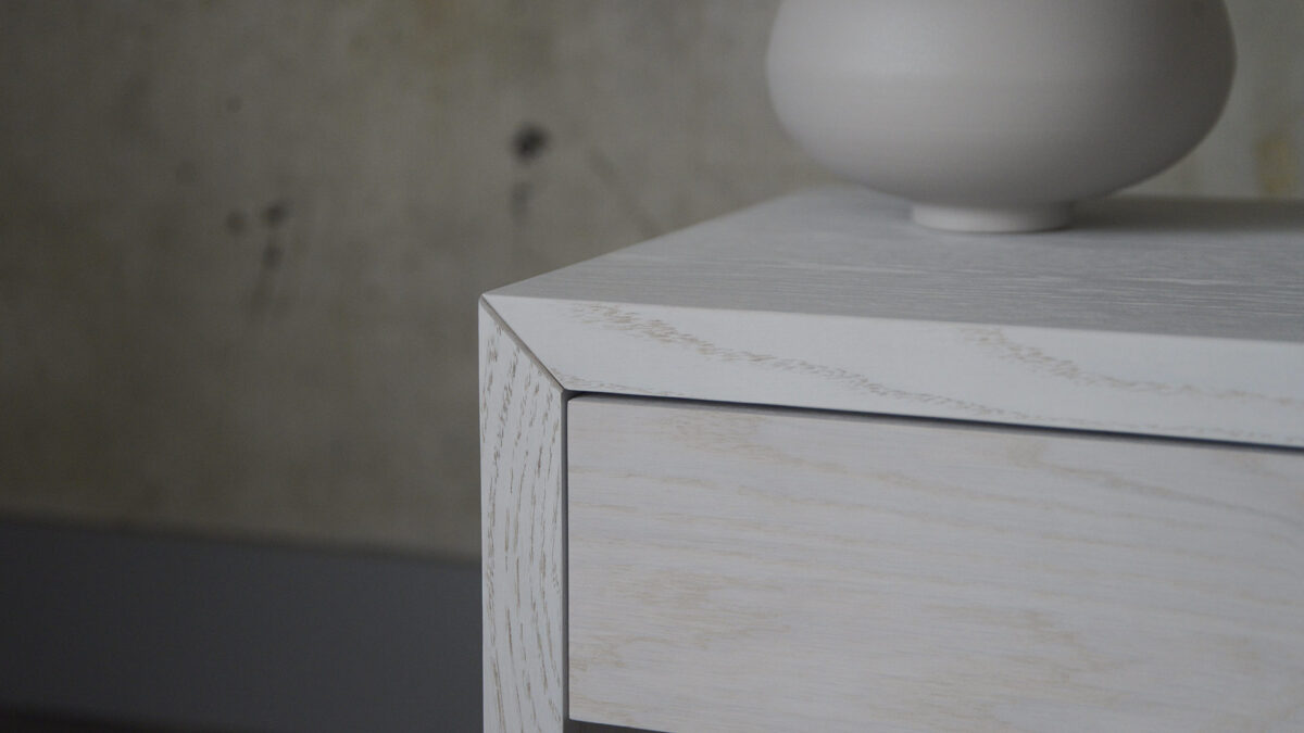 Close-up image of a small bedside table, showing the grain of the timber through a pure white stain.
