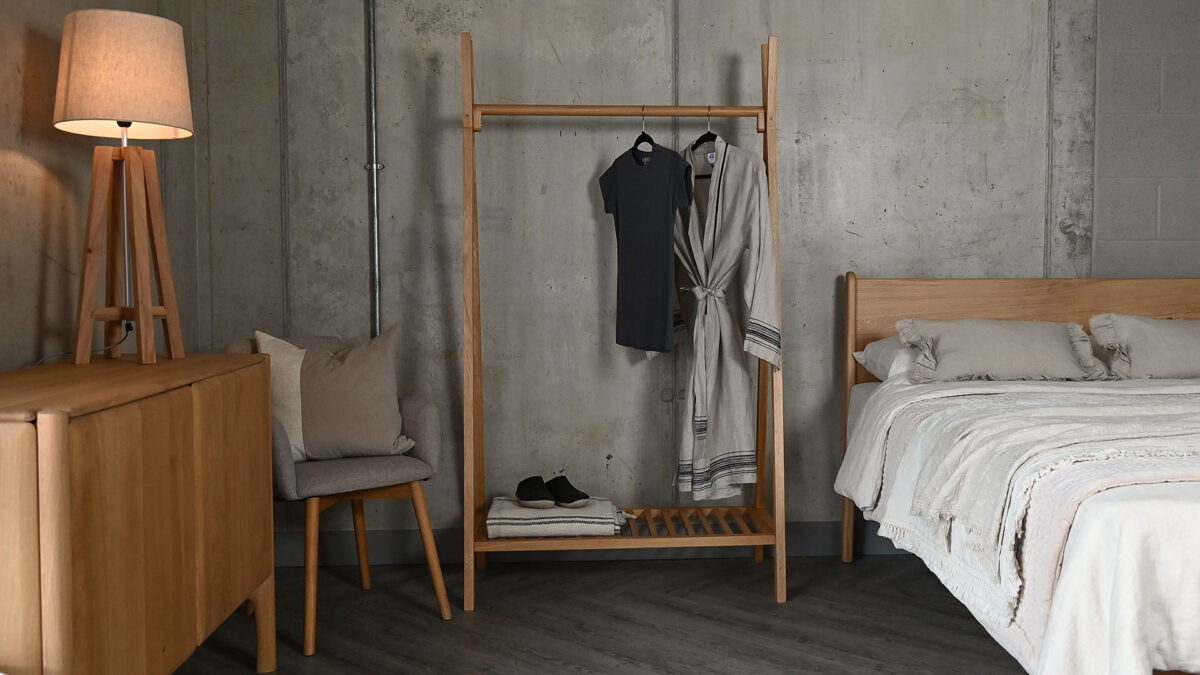 Solid oak wooden clothes rail with a grey t-shirt and cotton robe hanging from the rail, and a blanket and slippers stored on its lower shelf.