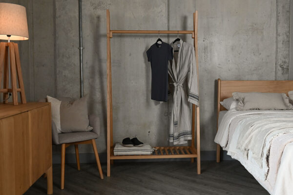 Solid oak wooden clothes rail with a grey t-shirt and cotton robe hanging from the rail, and a blanket and slippers stored on its lower shelf.